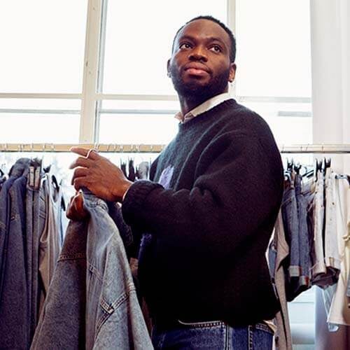 A male standing in front of a rail of clothes. Holding a few items of clothing, looking off camera.